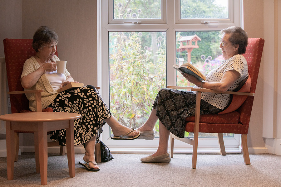 Winner of the South Tyneside Homes photography competition: two elderly women having a conversation in front of a care home window, one holds a cup of tea, while the other holds an open book